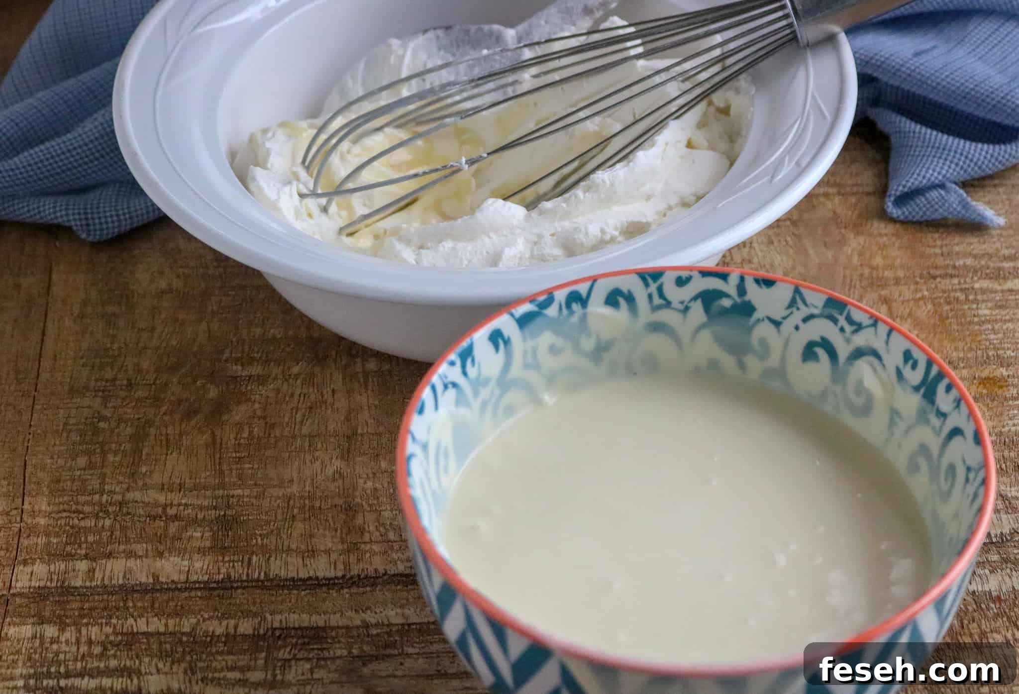 Thick heavy whipping cream in a white bowl with a whisk and sweetened condensed milk in a teal patterned bowl on a wood table next to a blue kitchen towel
