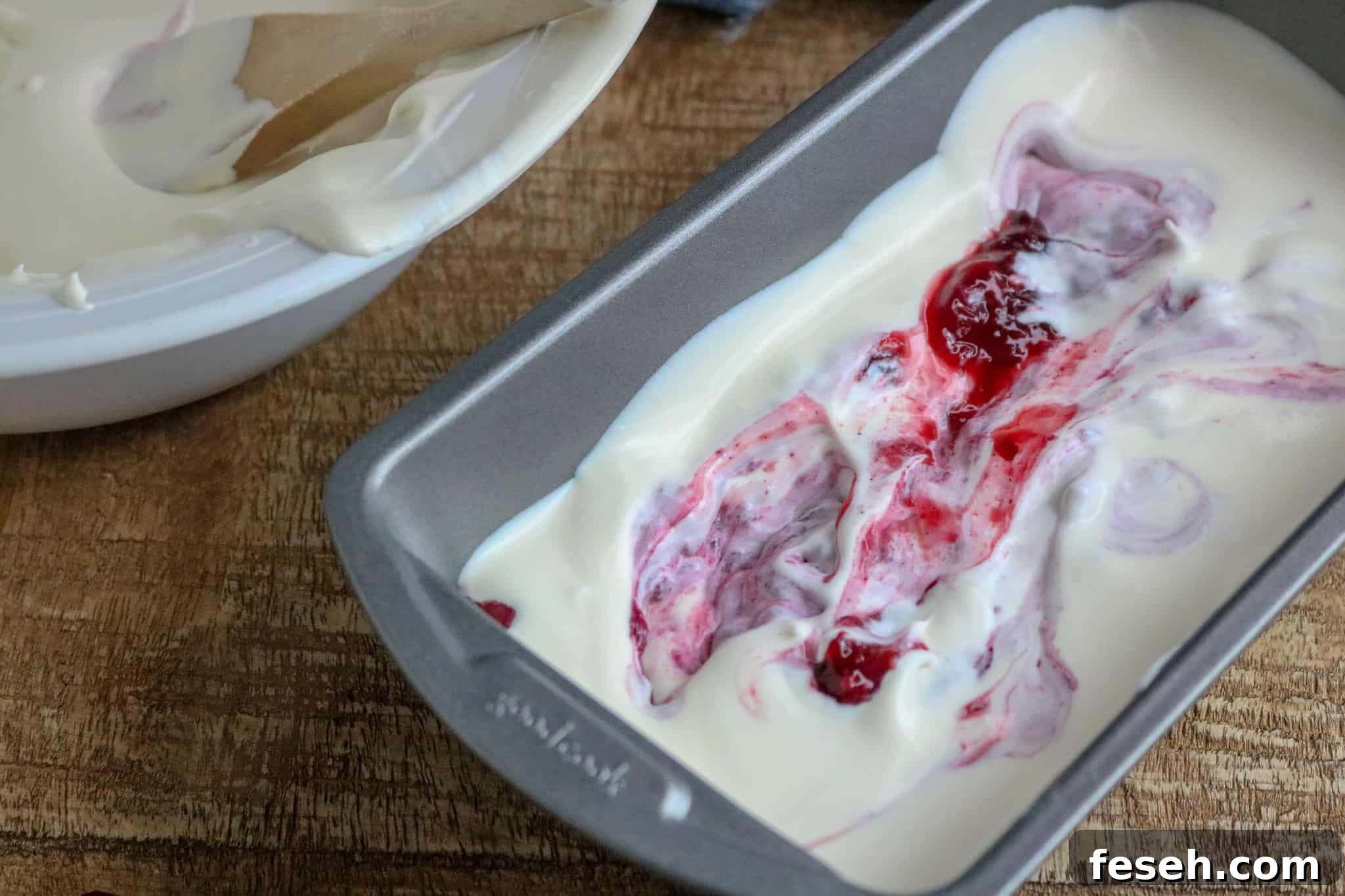 White bowl with No-Churn Ice Cream and wooden spoon next to a silver pan half-filled with No-Churn Ice Cream with berry pie filling swirled on top