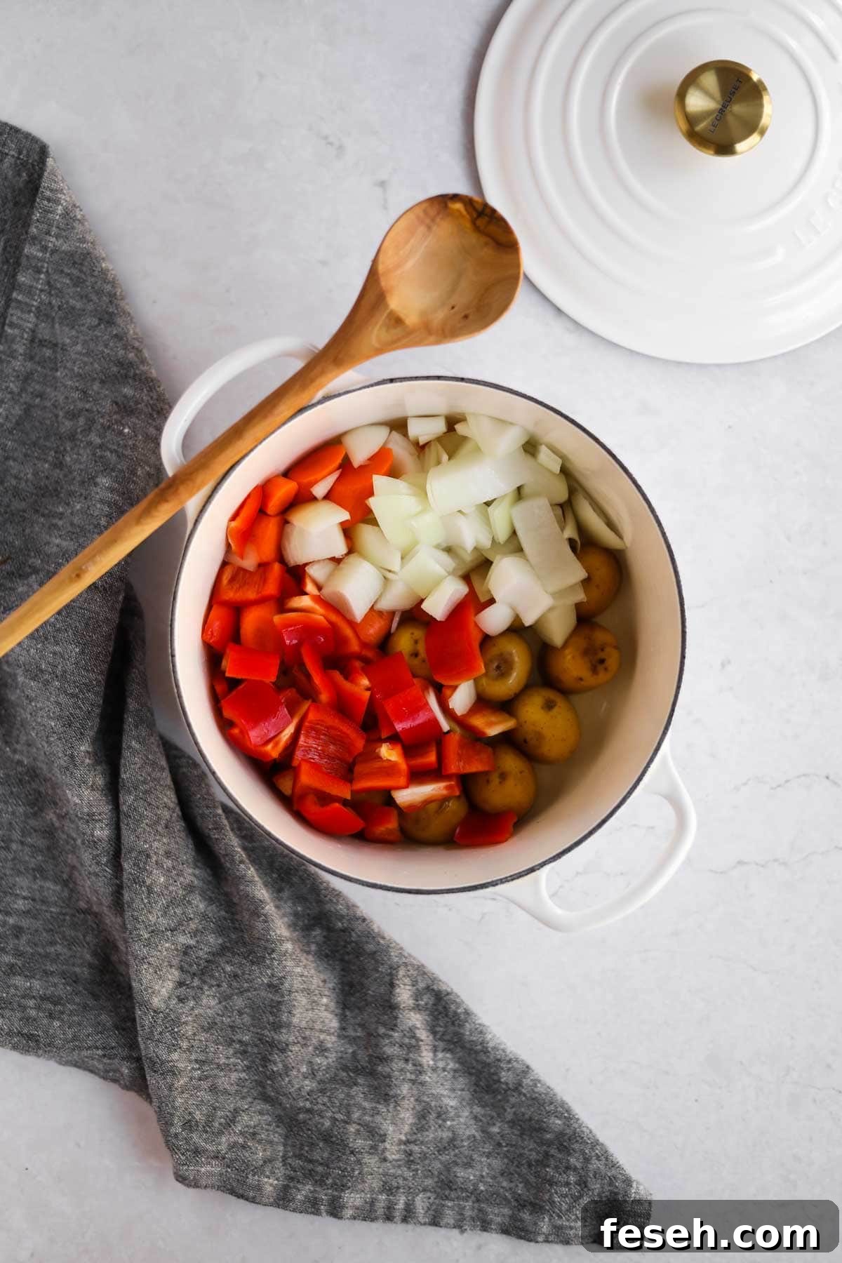 Overhead shot of onion, potatoes, bell peppers in white Le Creuset dutch oven with wooden spoon