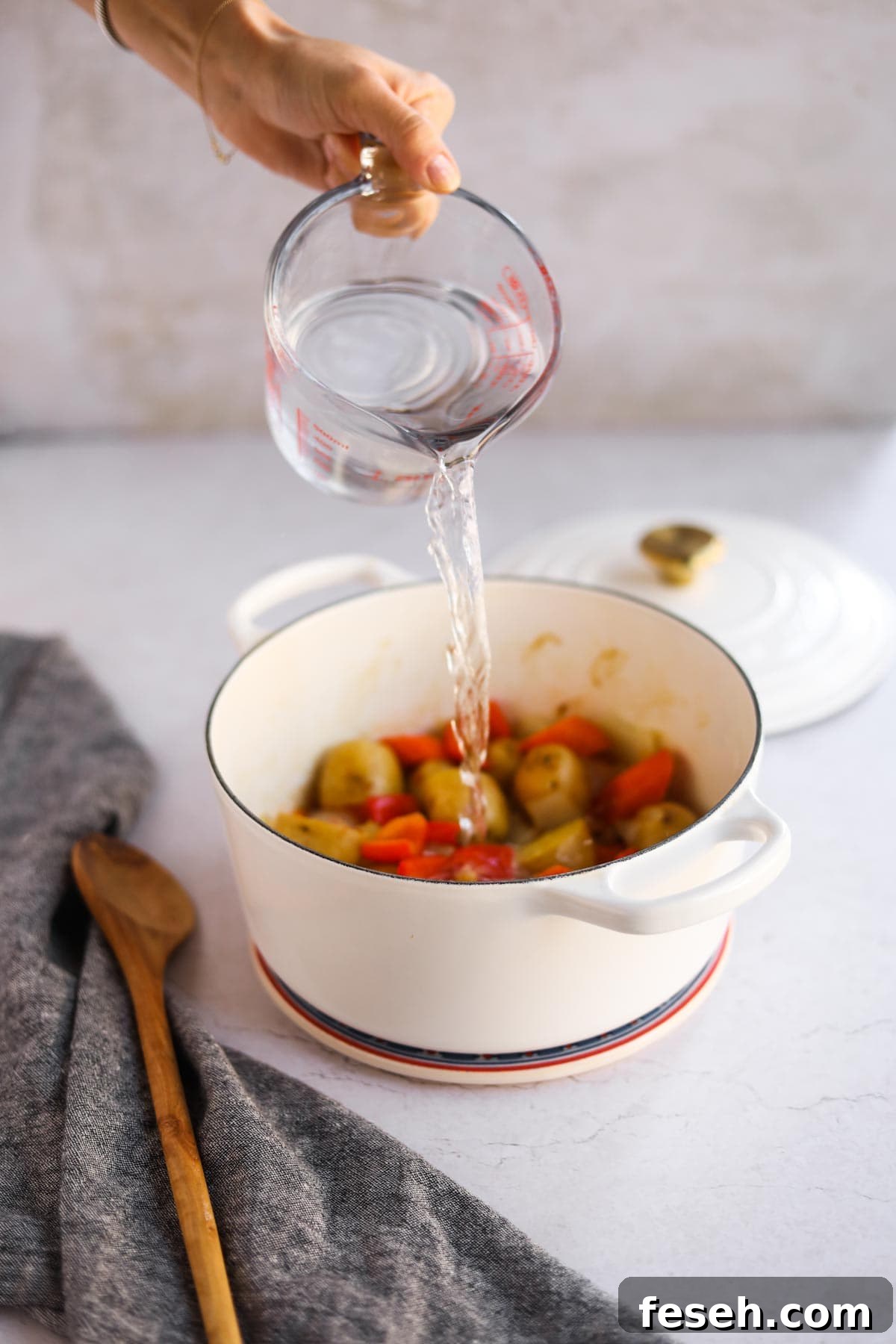 Measuring cup pouring water on onion, potatoes, and bell peppers in white Le Creuset dutch oven with wooden spoon