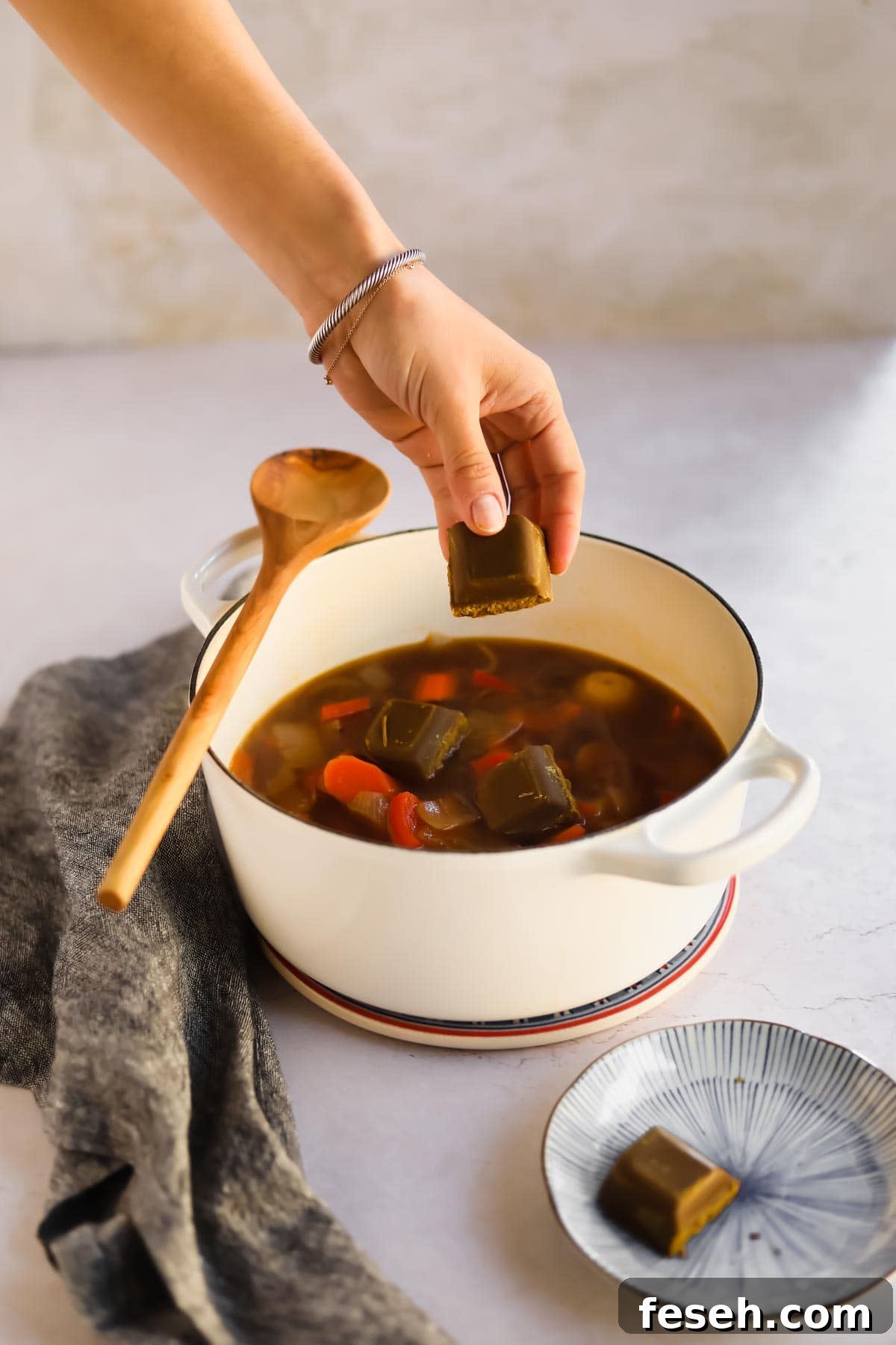 Placing Golden Curry Rou in white Le Creuset dutch oven containing on onion, potatoes, and bell peppers in with wooden spoon