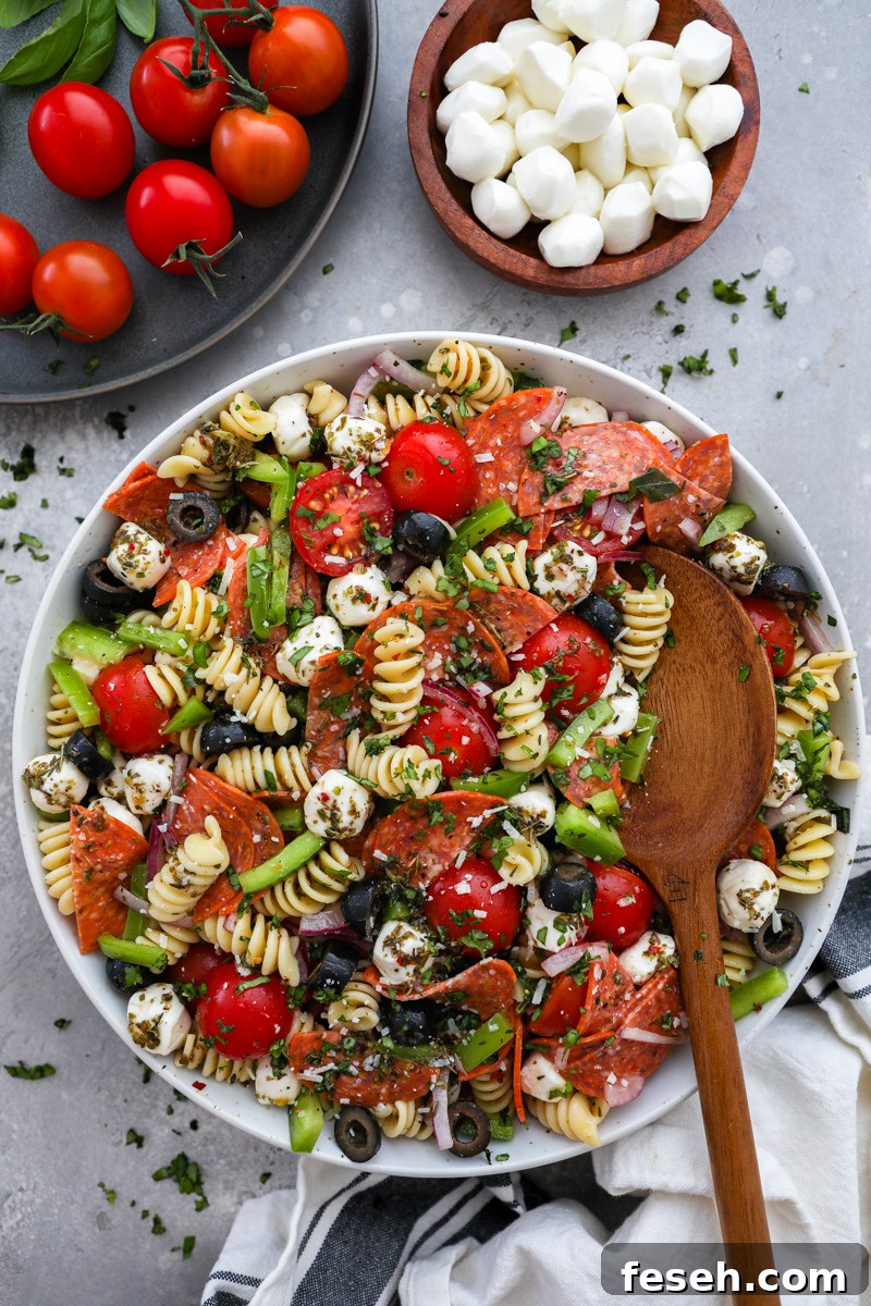 A large white serving bowl filled with pizza pasta salad surrounded by a small bowl of fresh mozzarella pearls, vine tomatoes and fresh basil