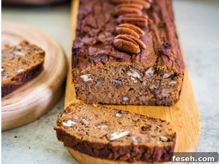 A loaf of keto pumpkin bread sits on a wooden board, topped with whole pecans. Two slices are cut, with one resting invitingly on a nearby plate.