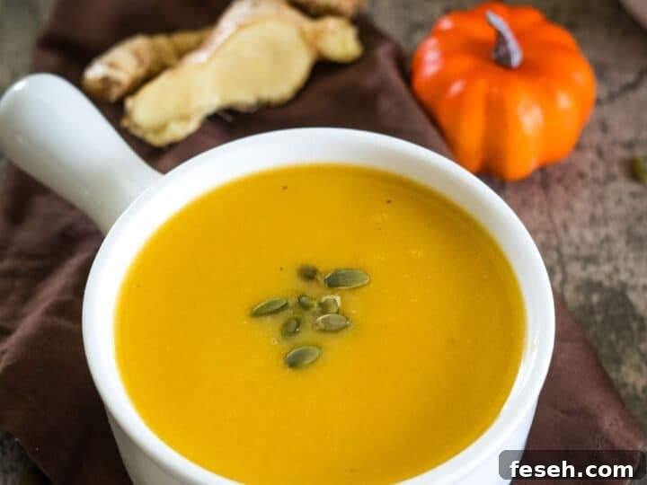 A bowl of pumpkin ginger soup garnished with pumpkin seeds, placed on a brown cloth beside ginger slices and a small decorative pumpkin.