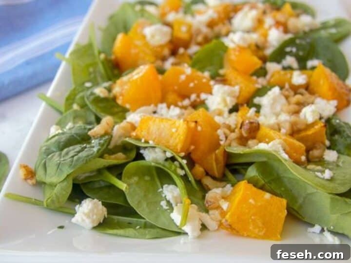 A plate of spinach salad topped with roasted pumpkin, walnuts, and crumbled feta cheese, served on a white square plate with a blue-striped napkin in the background.