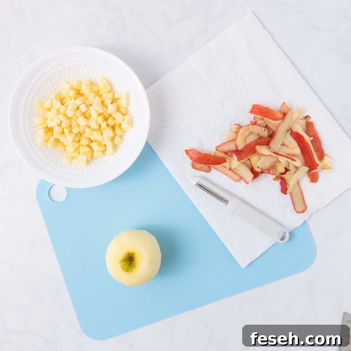 A peeled red apple on a blue chopping board, next to a bowl of chopped apple.