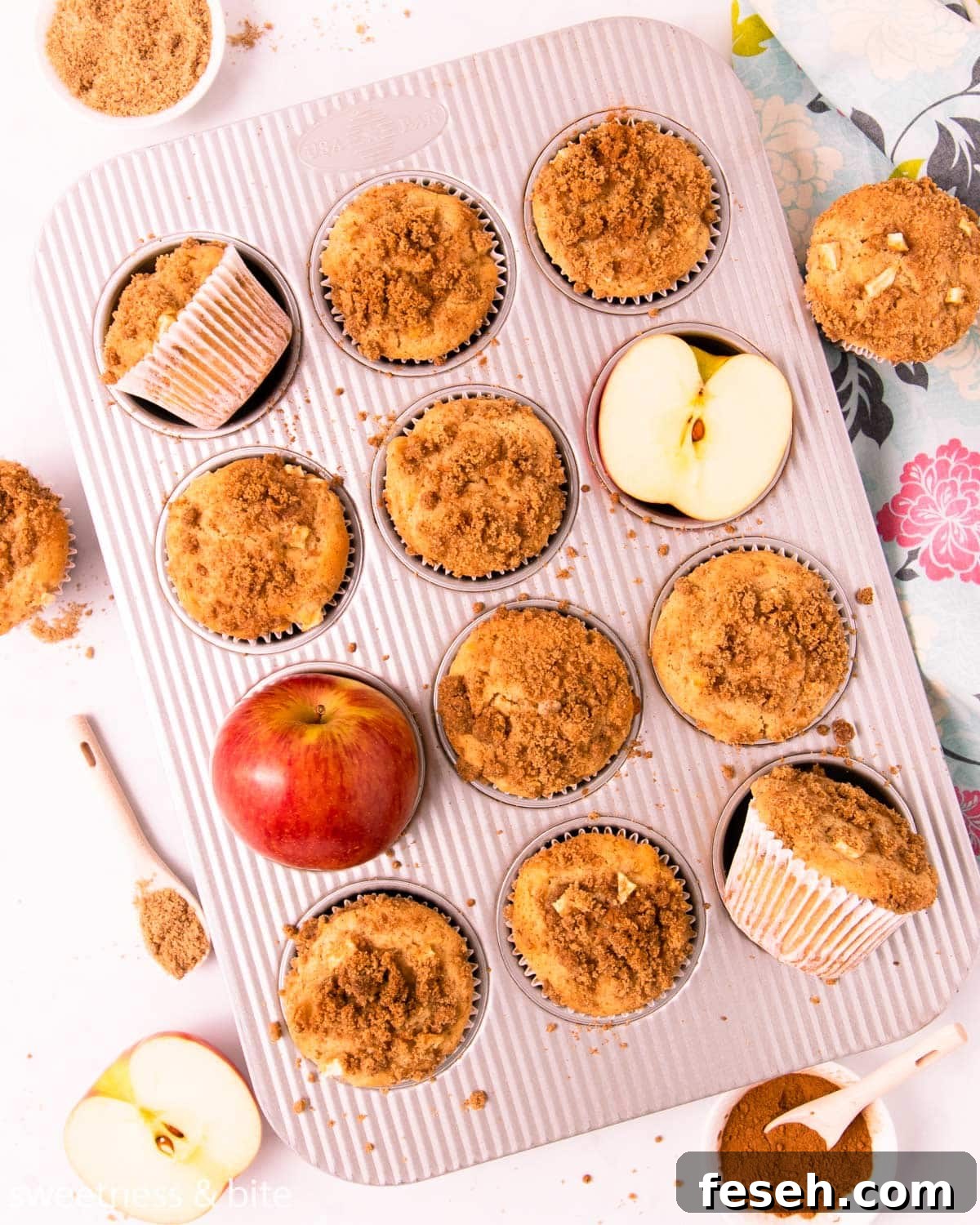 Gluten free apple muffins in a metal muffin pan, with a red apple and small dish of cinnamon streusel.