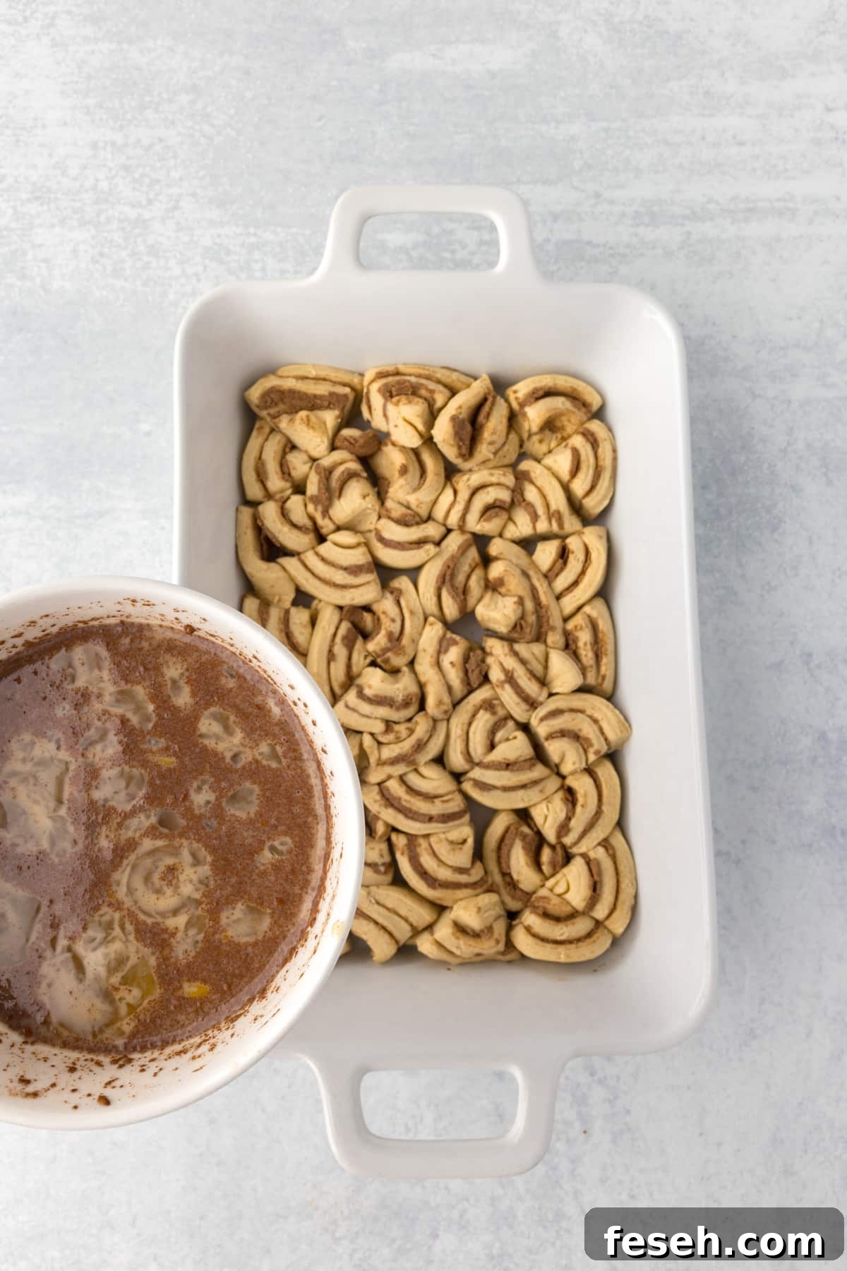 Pouring a custard mixture over a baking dish filled with cinnamon roll pieces.