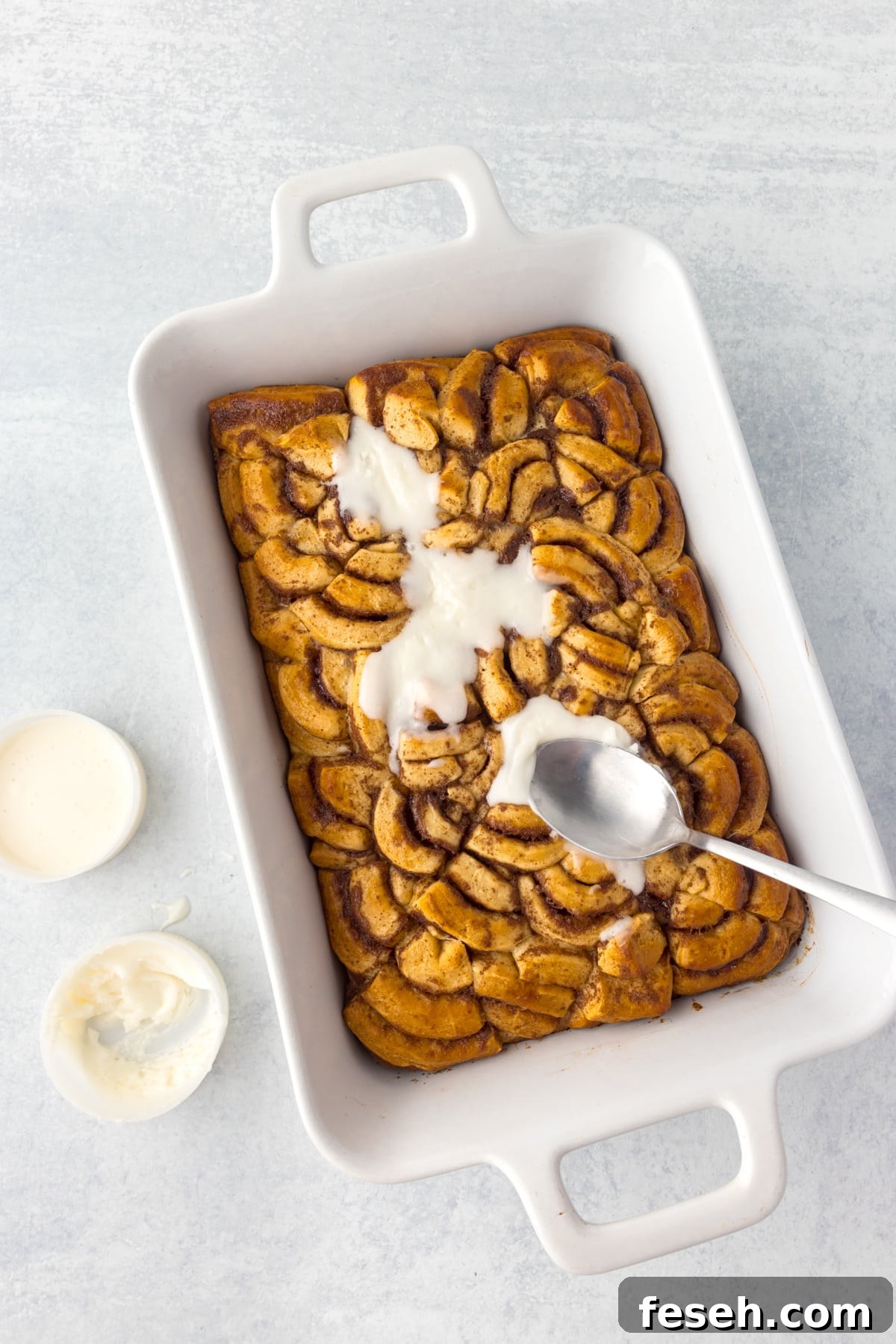 Spreading glaze on a baking dish with baked cinnamon roll pieces.