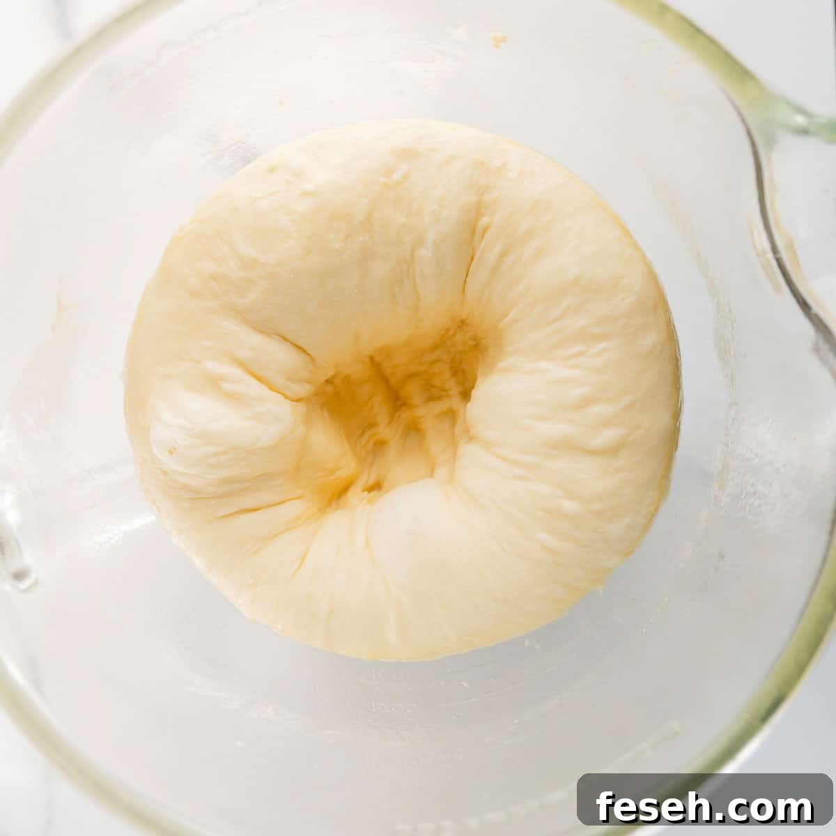 A close-up view of bread dough in a glass bowl, showing a dimple in the center.