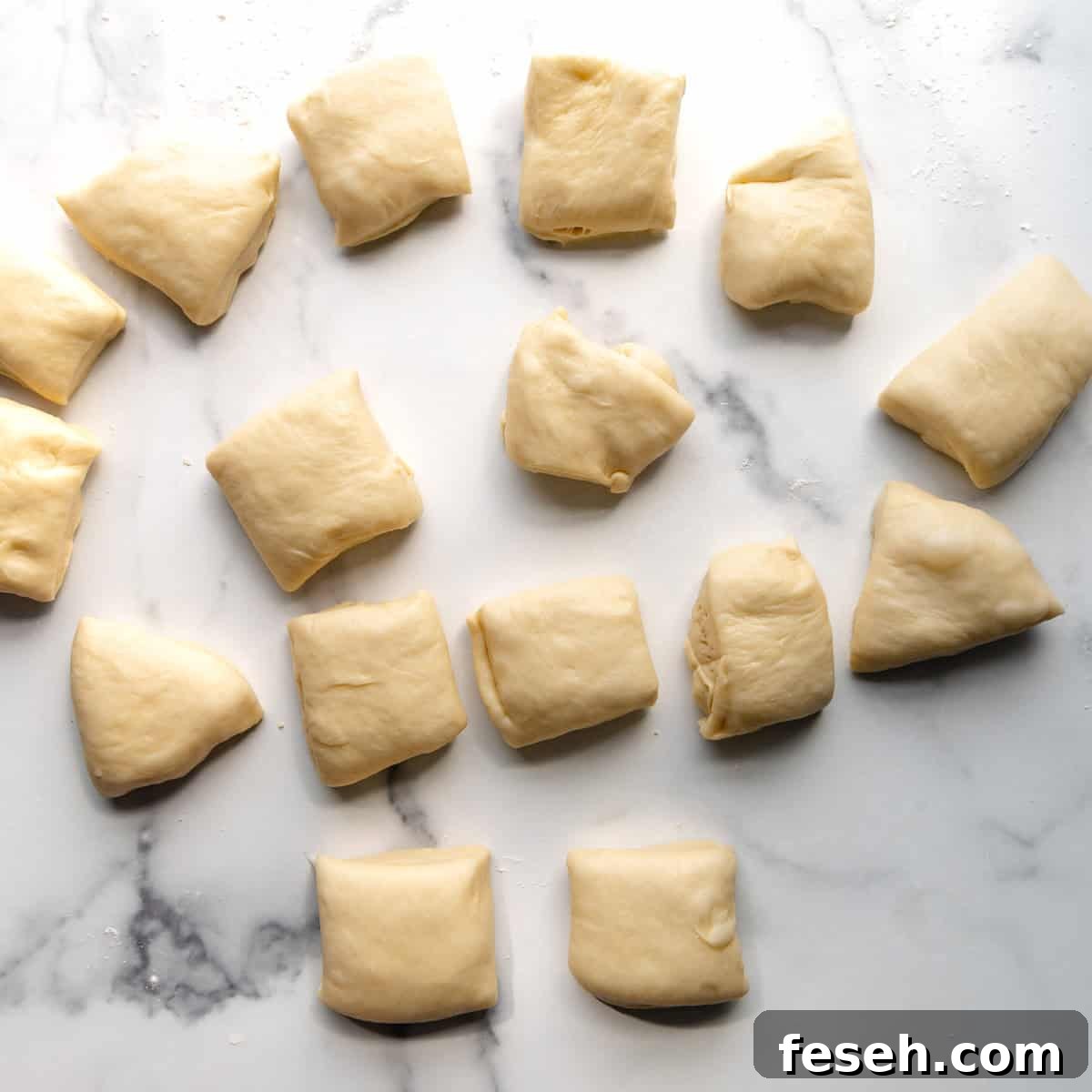 Close-up of 15 pieces of raw dough on a white marble surface. The dough pieces are irregularly shaped and spaced.