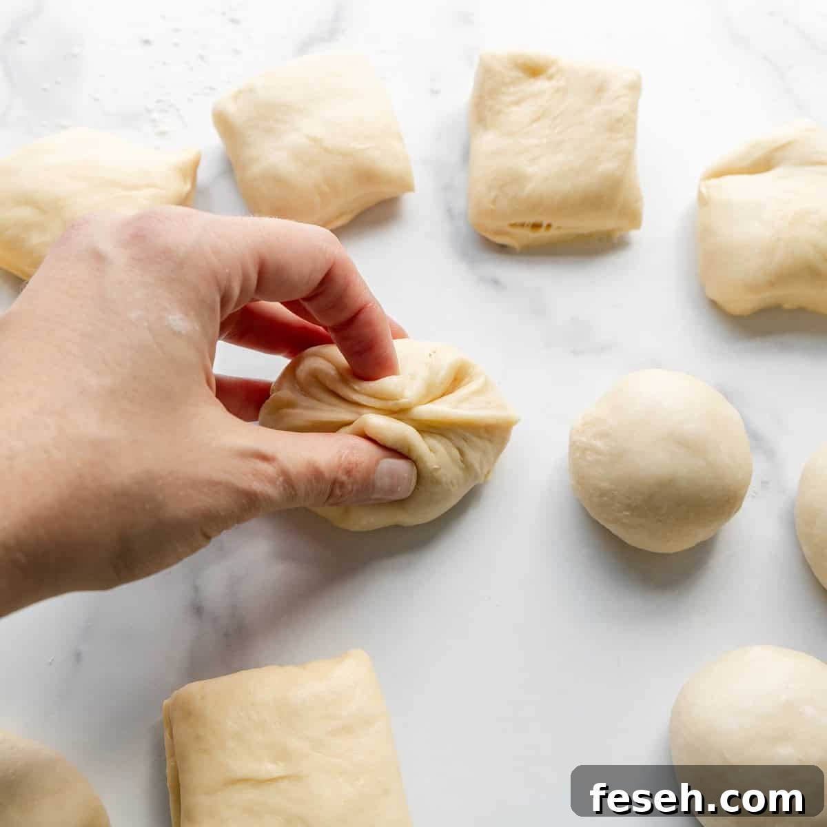 A hand folding a piece of dough on a white surface. Other pieces of dough, both square and round, are placed around.