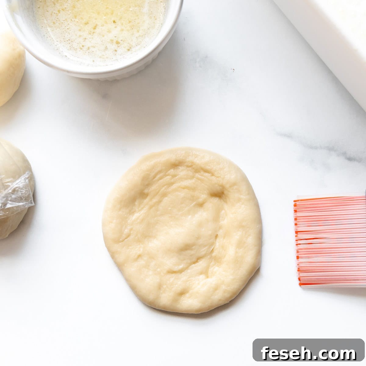 Flattened piece of dough on a white surface with a brush and bowl of partially mixed liquid nearby.