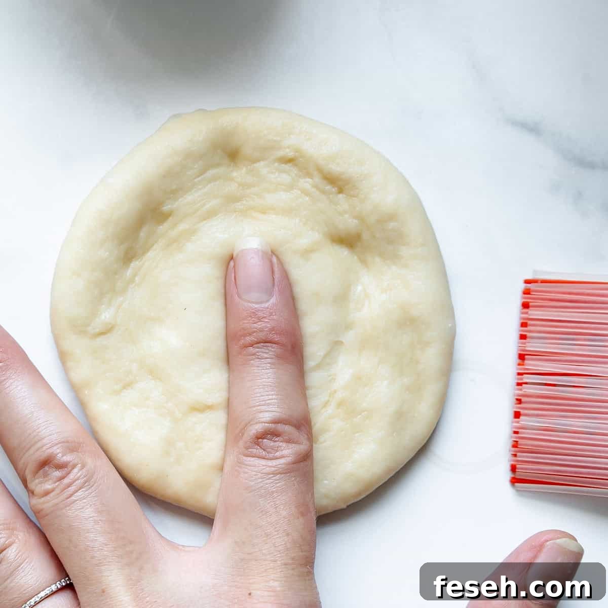 A person presses a finger into a small round piece of dough on a white surface next to an orange and white dough scraper.