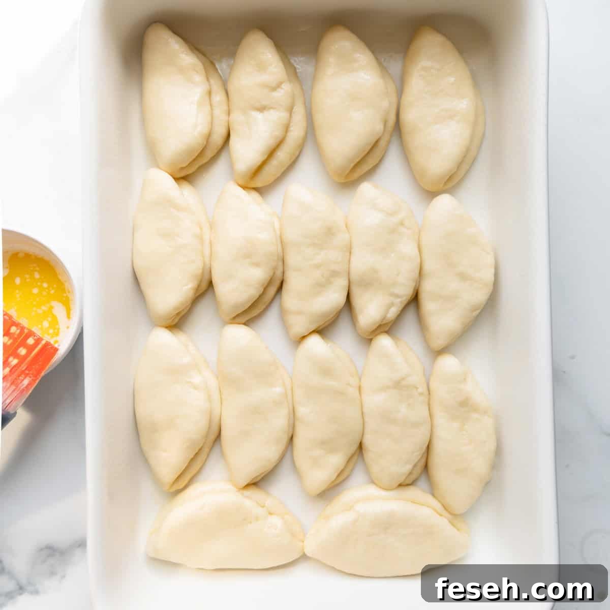 A baking dish filled with 16 uncooked dough pieces arranged in rows, near a bowl of melted butter with a brush.