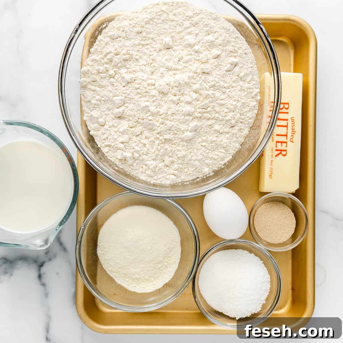 A baking tray with a glass of milk, a bowl of flour, a stick of butter, an egg, and smaller bowls containing sugar, yeast, and powdered milk.