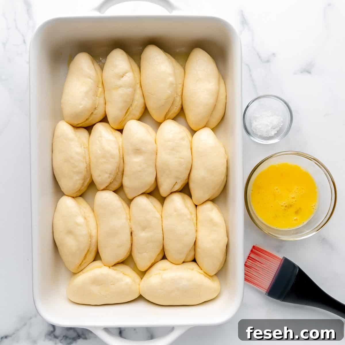 A baking dish with dough pieces arranged neatly. Beside it are a bowl of whisked egg, a brush, and a small bowl of salt.