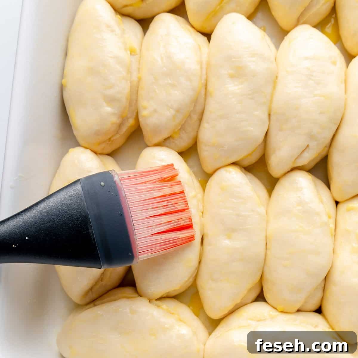 A close-up of a person using a pastry brush to apply egg wash onto several unbaked, dough-covered pastries arranged in a baking tray.