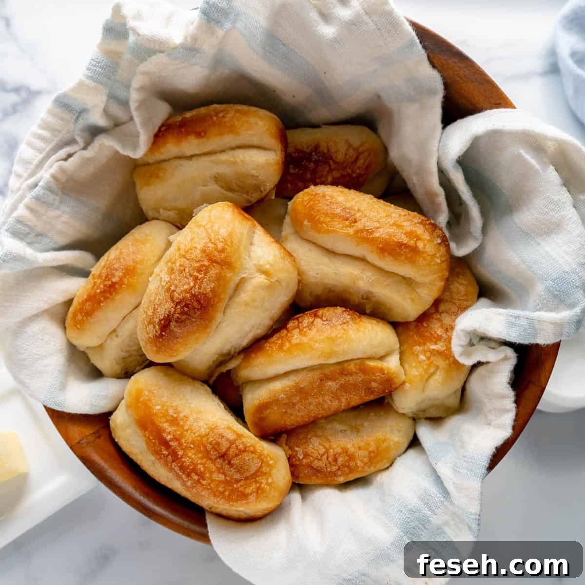 A bowl lined with a white cloth holds several freshly baked, golden-brown yeast rolls on a marble surface.