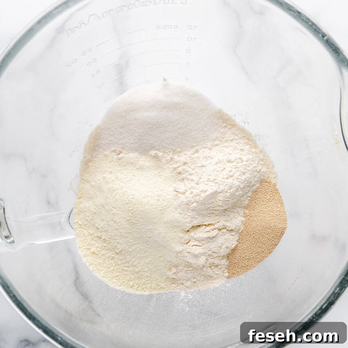 A glass mixing bowl containing dry baking ingredients including flour, sugar, and yeast on a white marble surface.