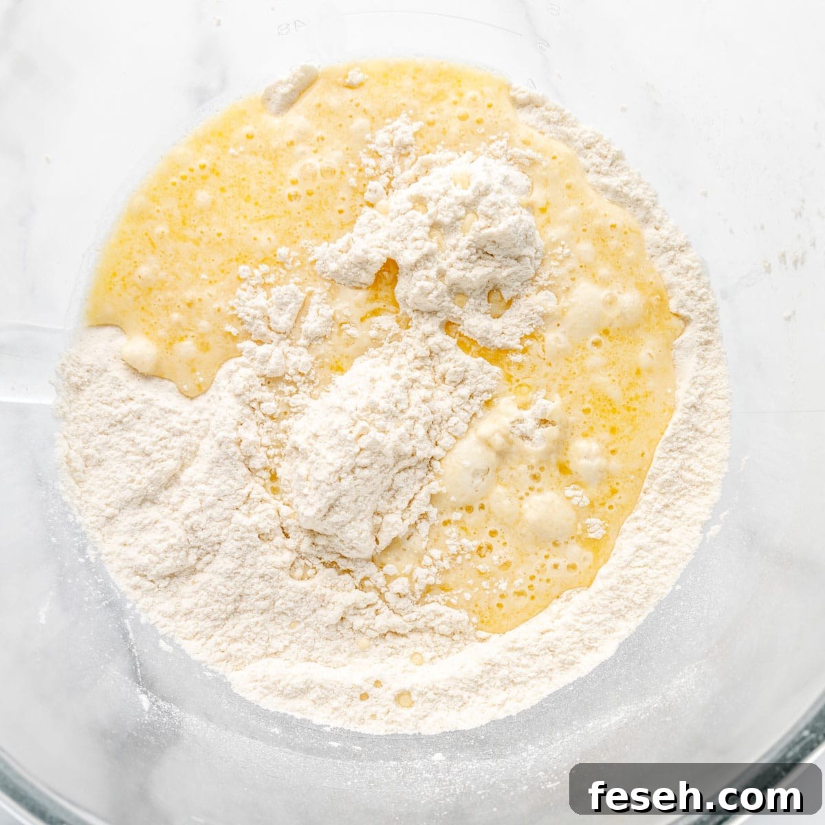 A mixing bowl containing flour and a yellow liquid mixture, likely egg and butter, in the process of being combined for baking.