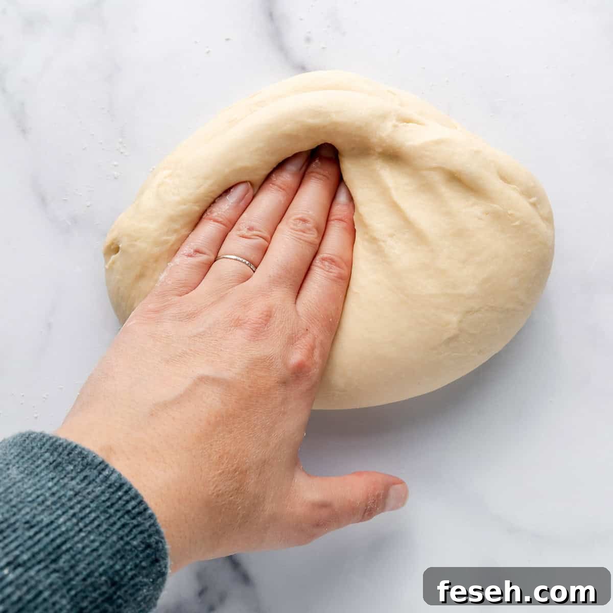 A hand with a ring presses down on a ball of dough on a marble surface.
