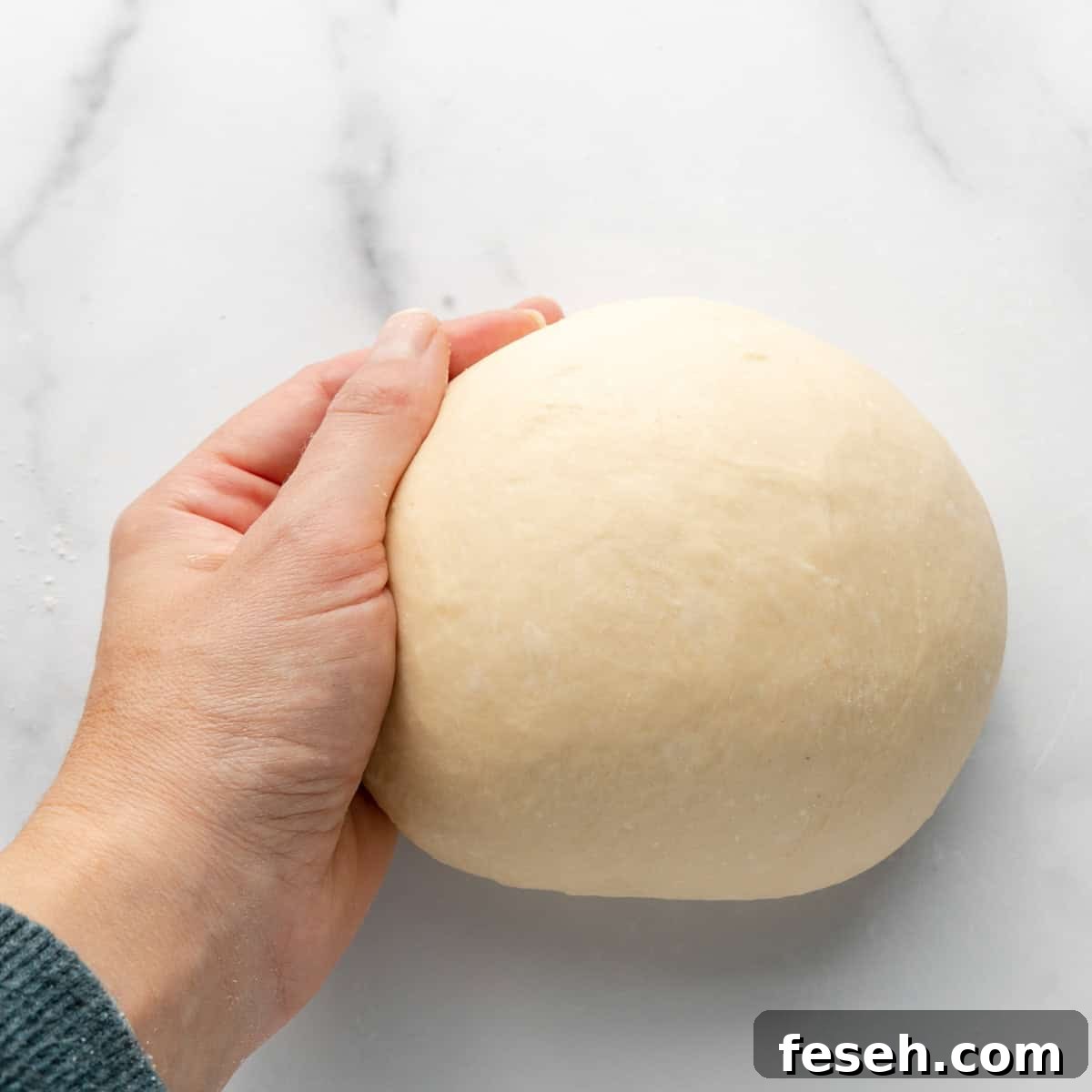 A hand holding a ball of dough on a white marble surface.