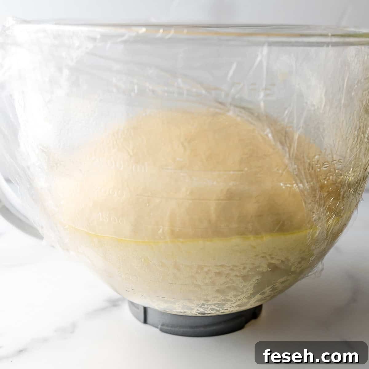 A glass bowl with dough rising inside, covered with plastic wrap. The bowl has measurement markings on the outside.