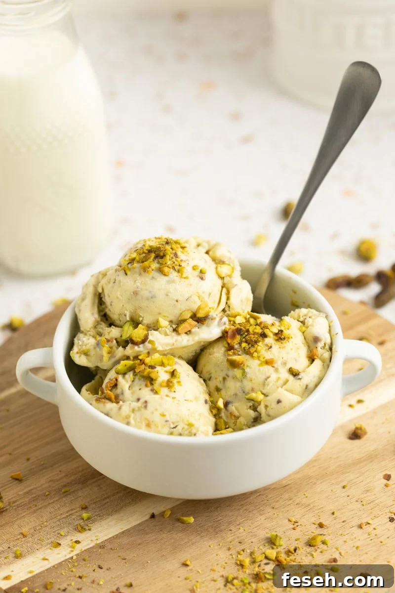 A white bowl of pistachio ice cream with a spoon in it and chopped pistachios laying around it