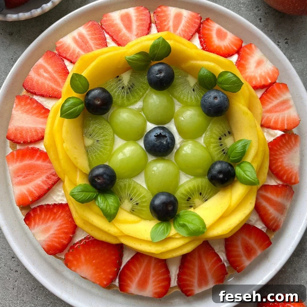A plate topped with a fruit pizza with neatly arranged fresh fruit with rings of sliced strawberries, mango slices, kiwi rounds, green grapes, blueberries, and basil leaves in the center.