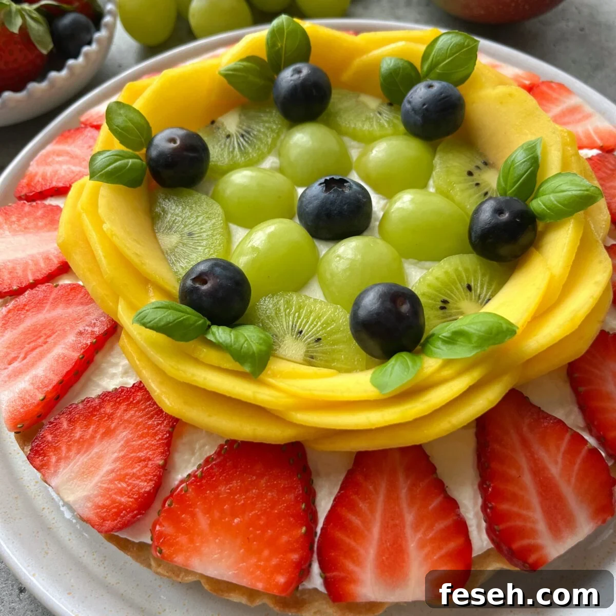 A fruit pizza topped with sliced strawberries, mango, kiwi, green grapes, blueberries, and small green leaves.