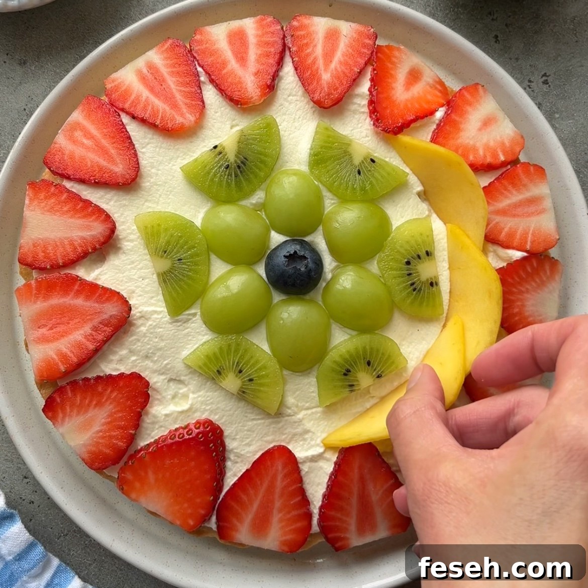 A hand places a slice of mango on a vibrant fruit pizza topped with whipped cream, strawberries, green grapes, kiwi slices, and a blueberry.