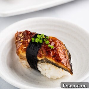 Close up of unagi sushi nigiri. Mound of rice topped with grilled Unagi, toasted sesame seeds, green onions and nori strip wrapped around.
