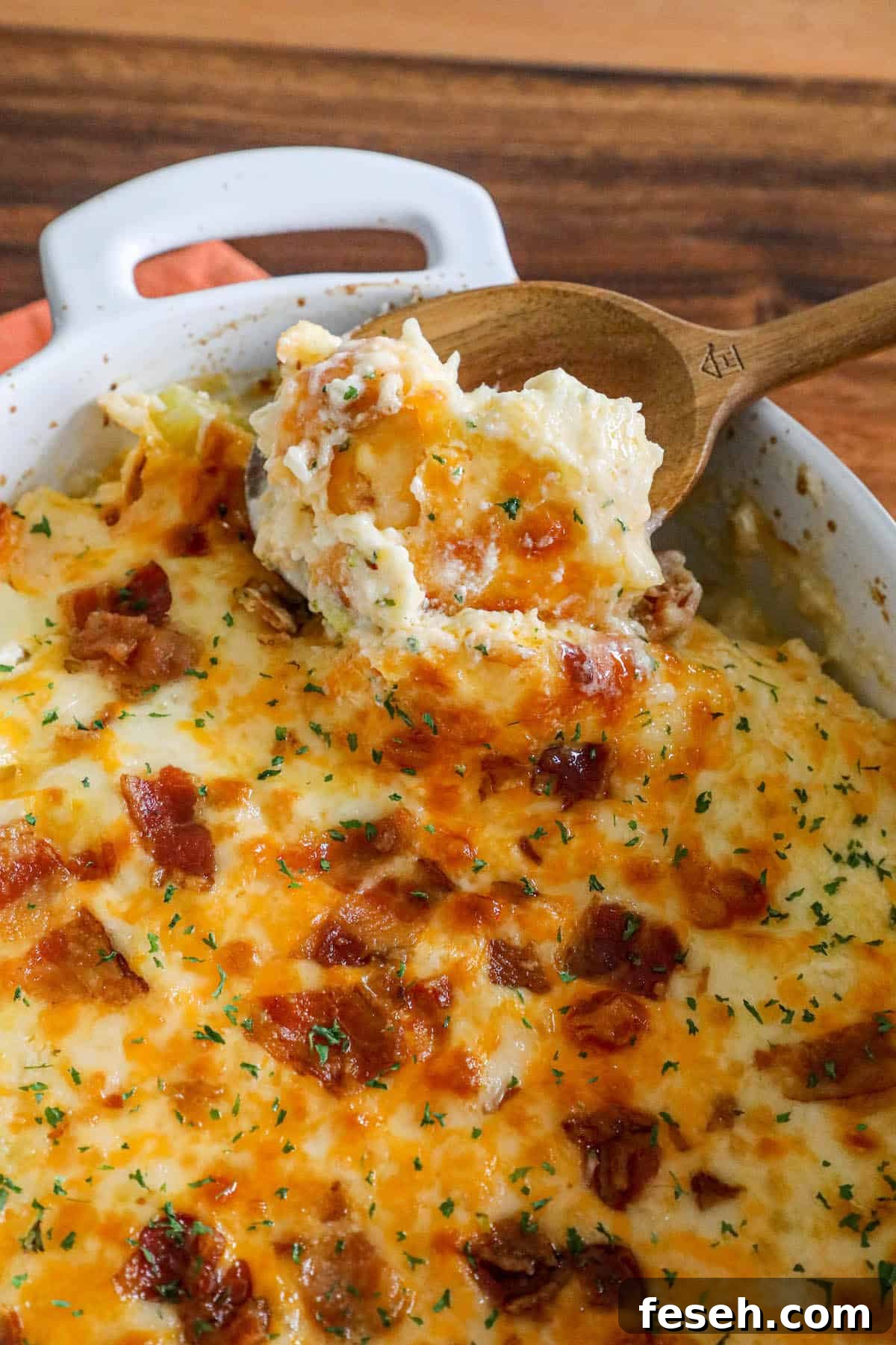 Cheesy mashed potatoes in a casserole dish with a wooden spoon, topped with bacon.
