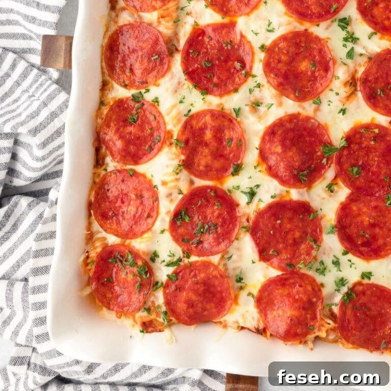 Close-up of Pizza Pasta Bake with melted cheese in a white dish, striped towel beside.