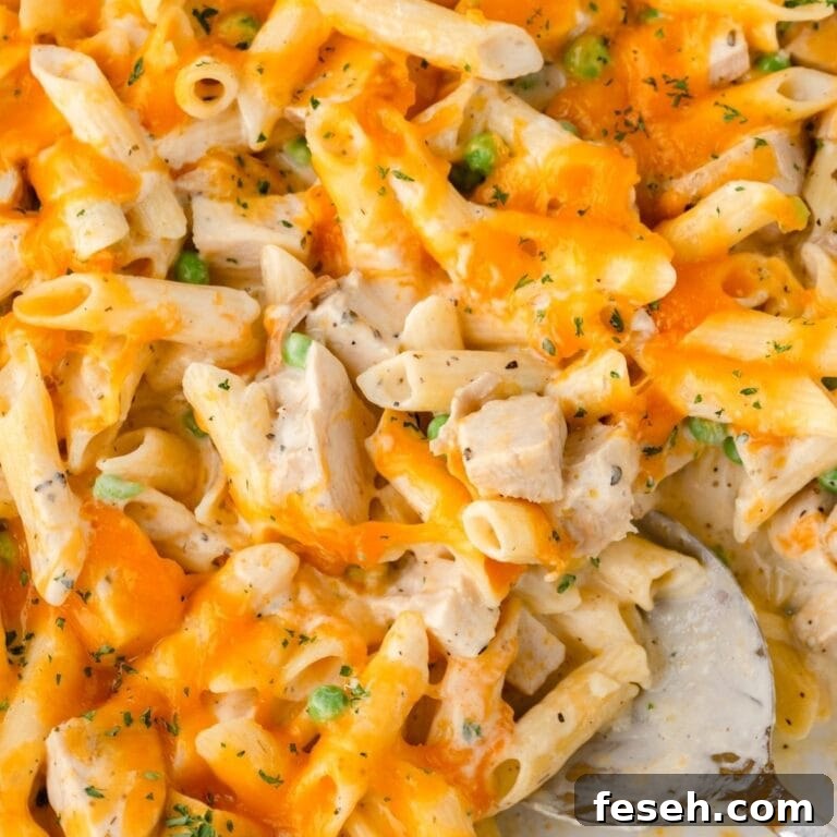 Creamy chicken pasta bake with penne, cheddar, green peas, herbs, and a silver serving spoon.