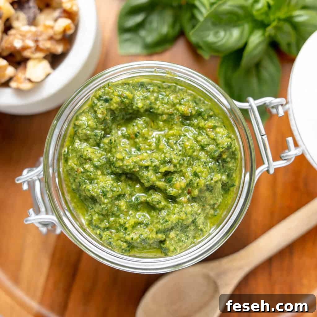 A glass jar filled with green pesto sits on a wooden surface, surrounded by fresh basil leaves, walnuts, and a wooden spoon.