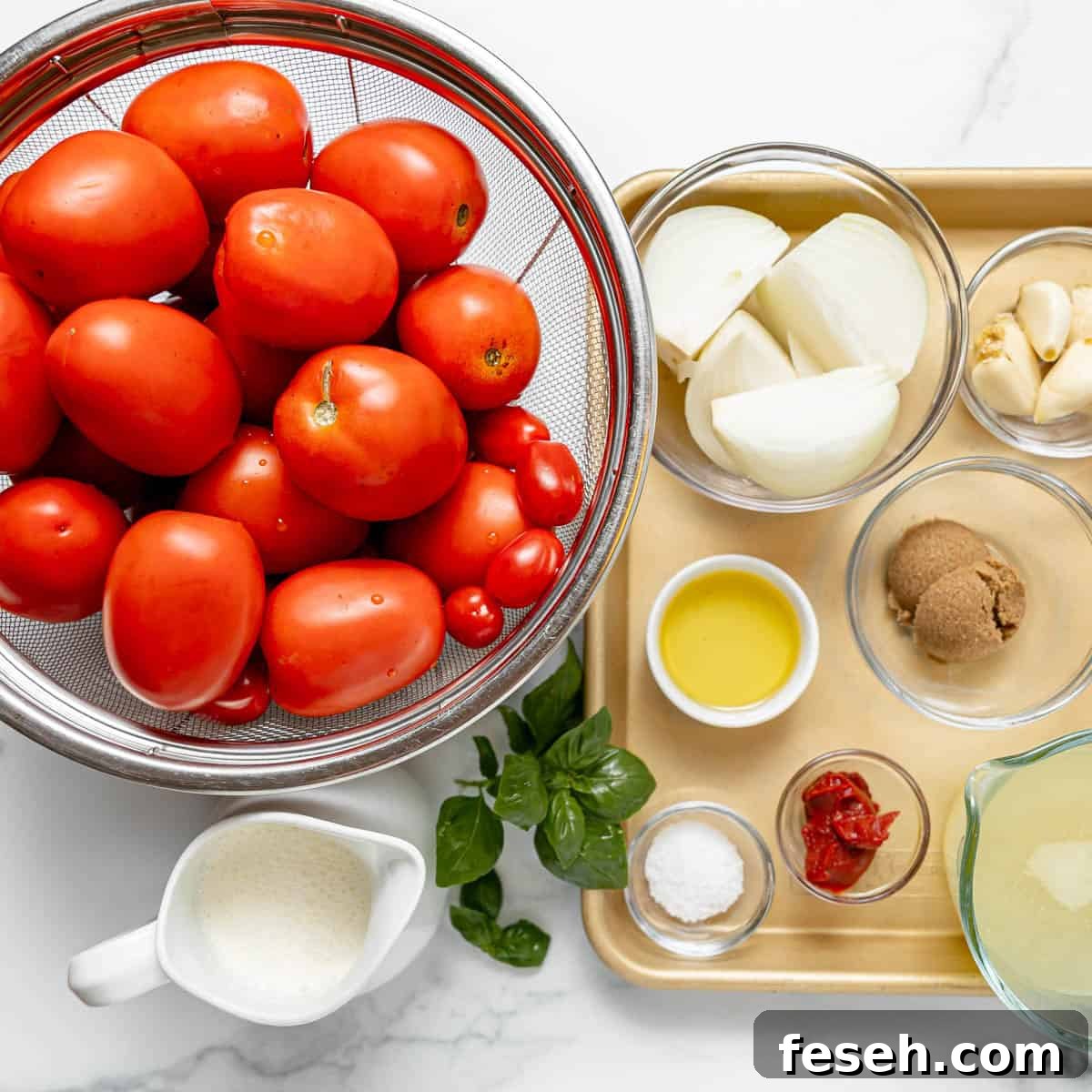 A colander of tomatoes, fresh basil, onion, garlic, olive oil, brown sugar, tomato paste, salt, broth, and cream arranged on a white countertop and a tray.