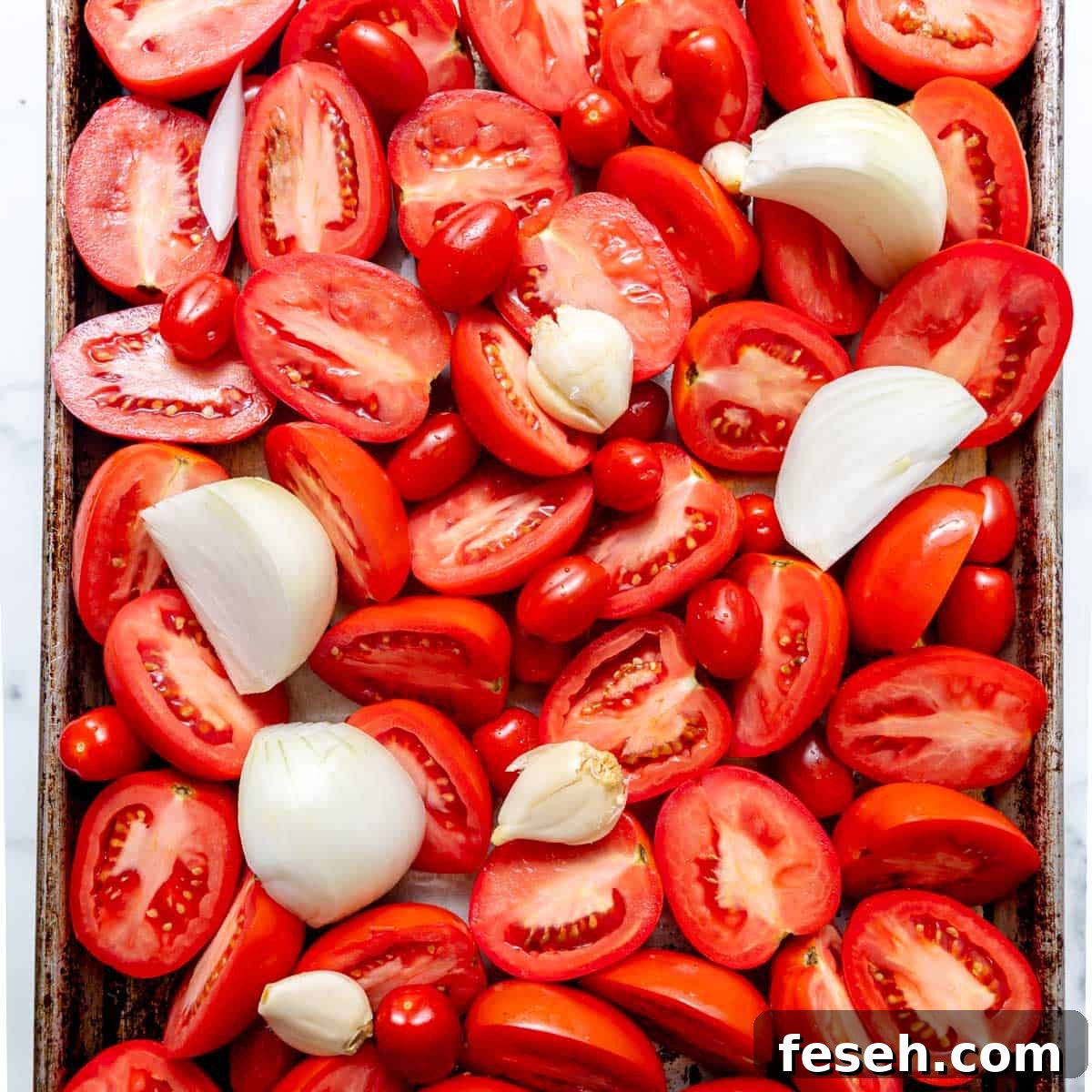 A baking tray filled with halved tomatoes, quartered onions, and whole garlic cloves, ready for roasting.