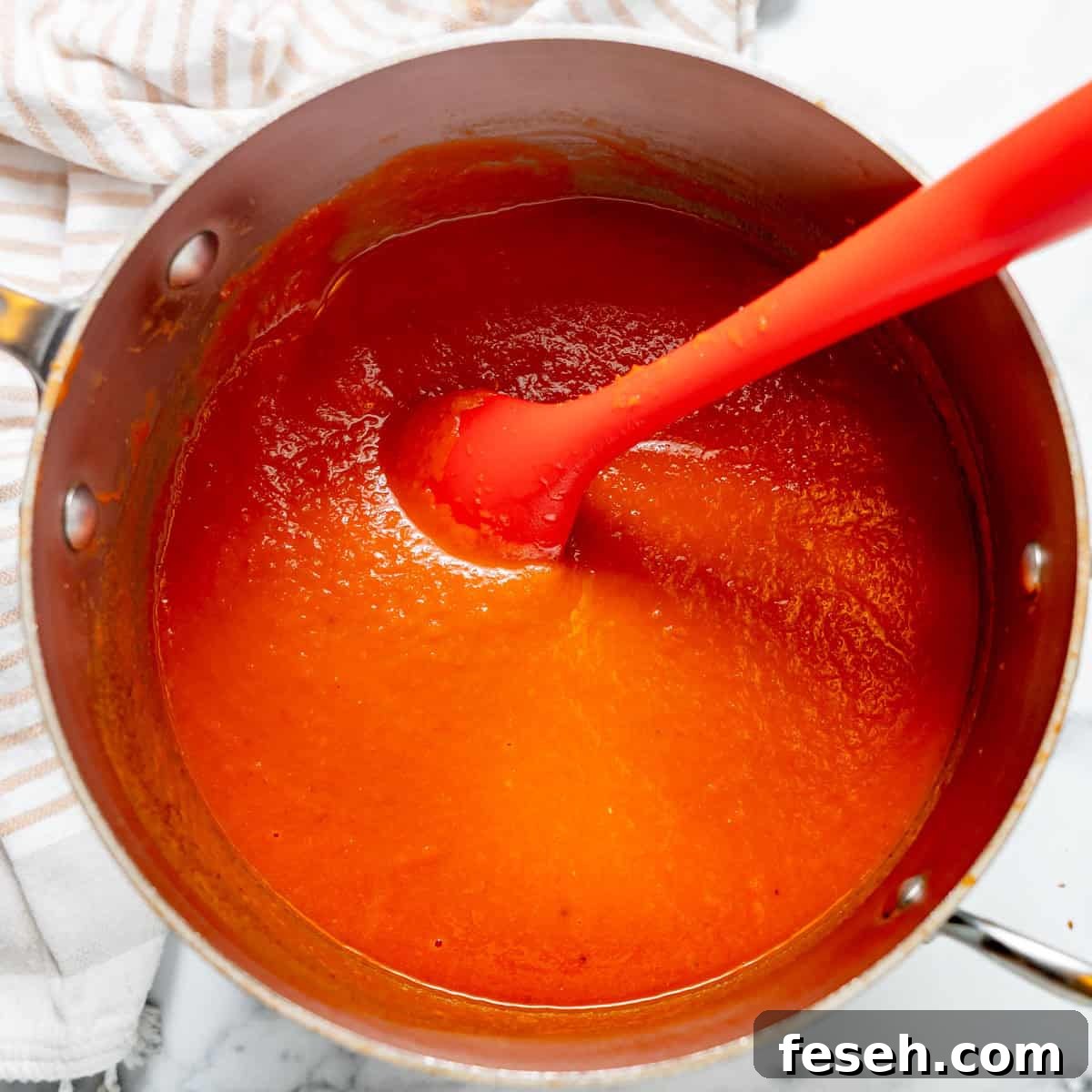 A pot of smooth, red tomato soup being stirred with a red silicone spatula on a marble surface next to a striped dish towel.