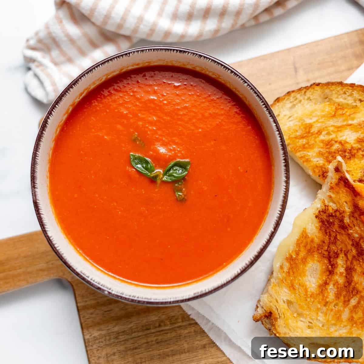 A bowl of tomato soup garnished with basil leaves next to a grilled cheese sandwich on a wooden board.