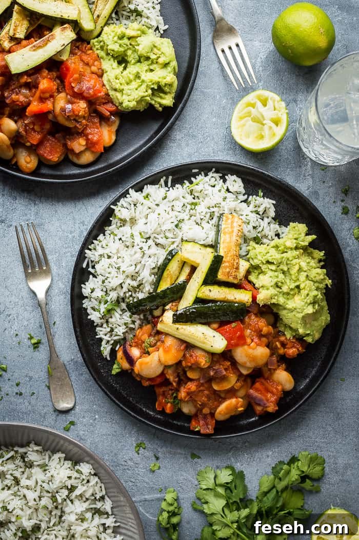 Two black plates of vegan lentil, vegetable and butter bean chilli, coriander rice, guacamole and roasted courgette on a grey background.