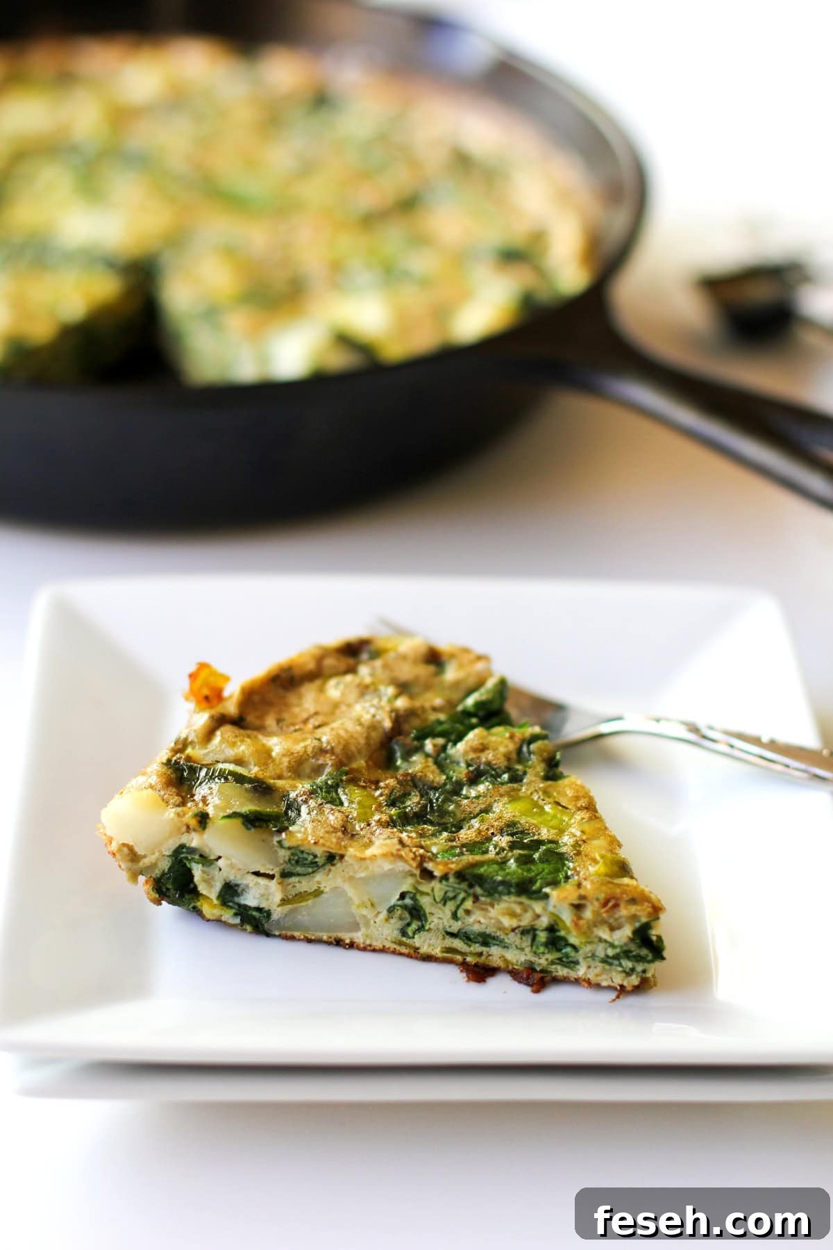 Slice of frittata on a white square plate with the rest of the frittata in a cast iron skillet in the background.