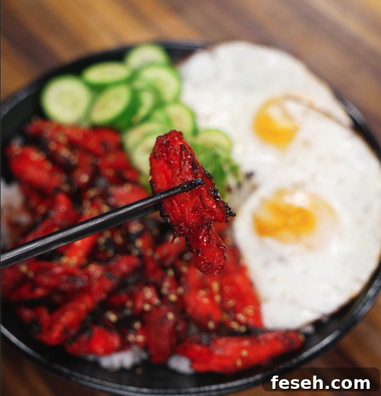 Close-up of a piece of glazed Chicken Tocino held with chopsticks over a bowl of rice, eggs, and vegetables. The sticky, sweet-savory exterior highlights the caramelization that makes tocino irresistible.