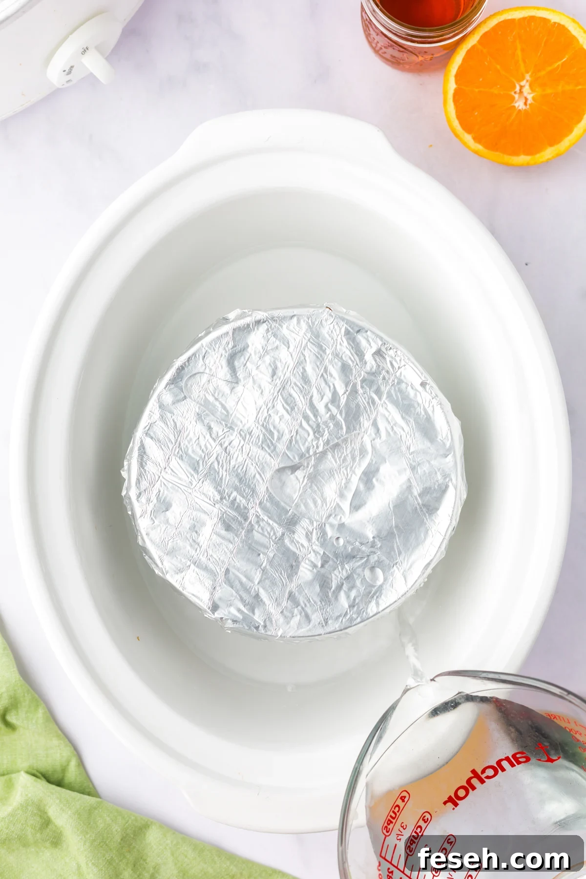 Foil-covered figgy pudding in a crockpot with water being poured around it.