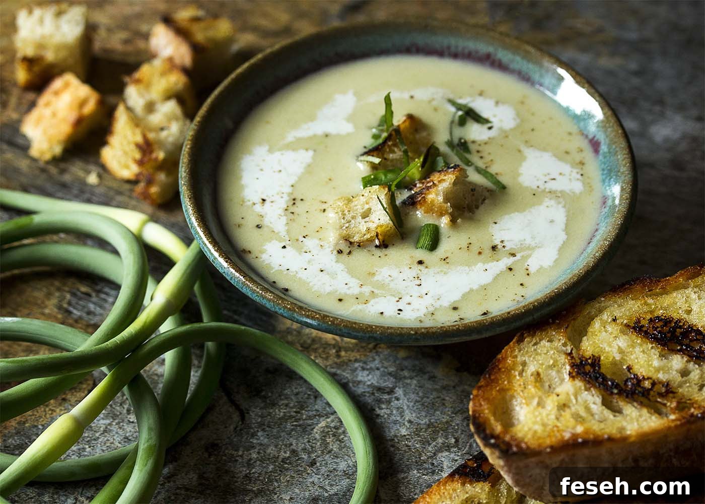 A bowl of cold garlic scape soup topped with garnishes. Grilled bread and garlic scapes scattered about.