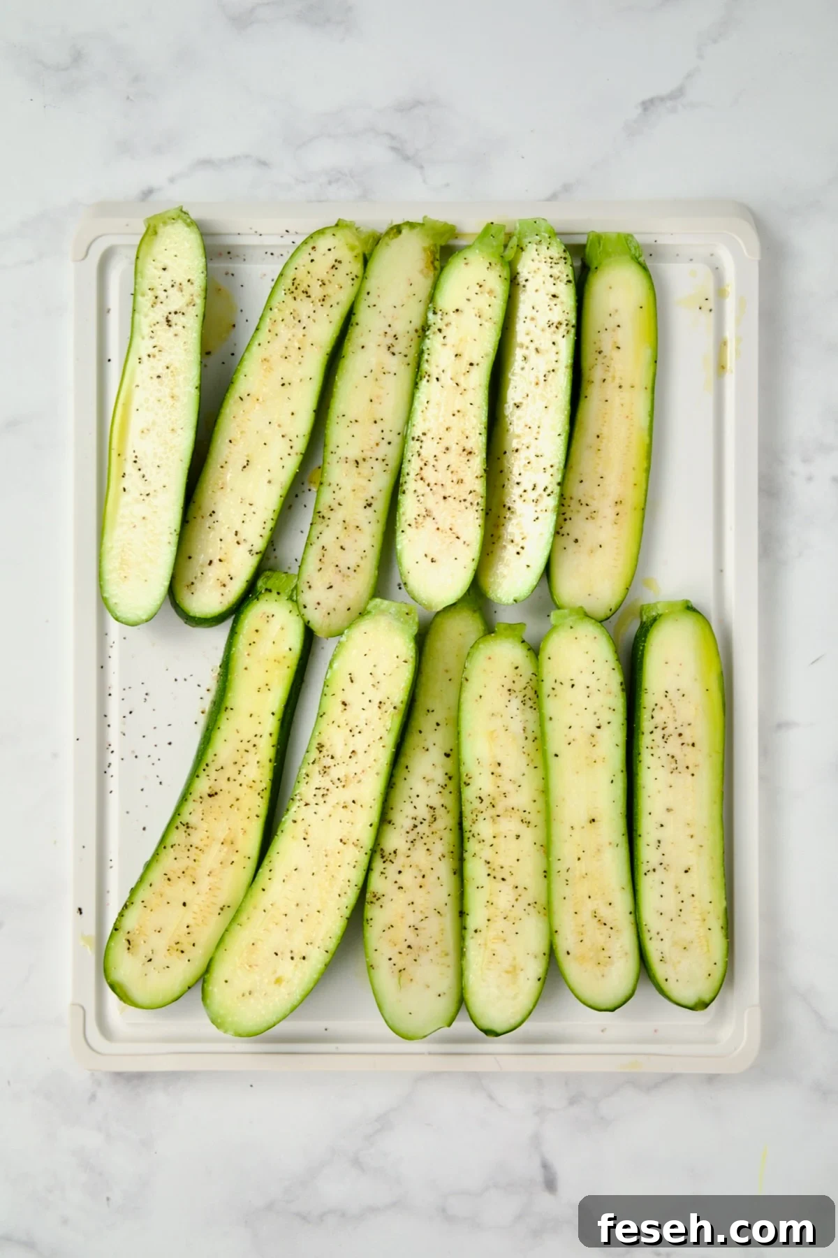 Sliced zucchini on white plastic cutting board seasoned with salt, pepper and drizzled with olive oil.