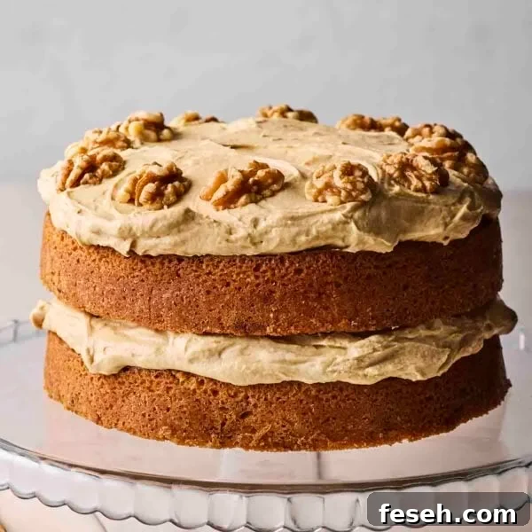 A side view of a classic Coffee and Walnut Cake with two layers of sponge and coffee buttercream and nuts on top, on a glass cake stand.