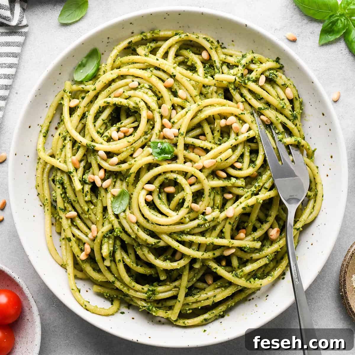 pesto pasta in a ceramic bowl with a silver fork, garnished with toasted pine nuts and fresh basil on a gray surface surrounded by basil, pine nuts, tomatoes and a striped towel.