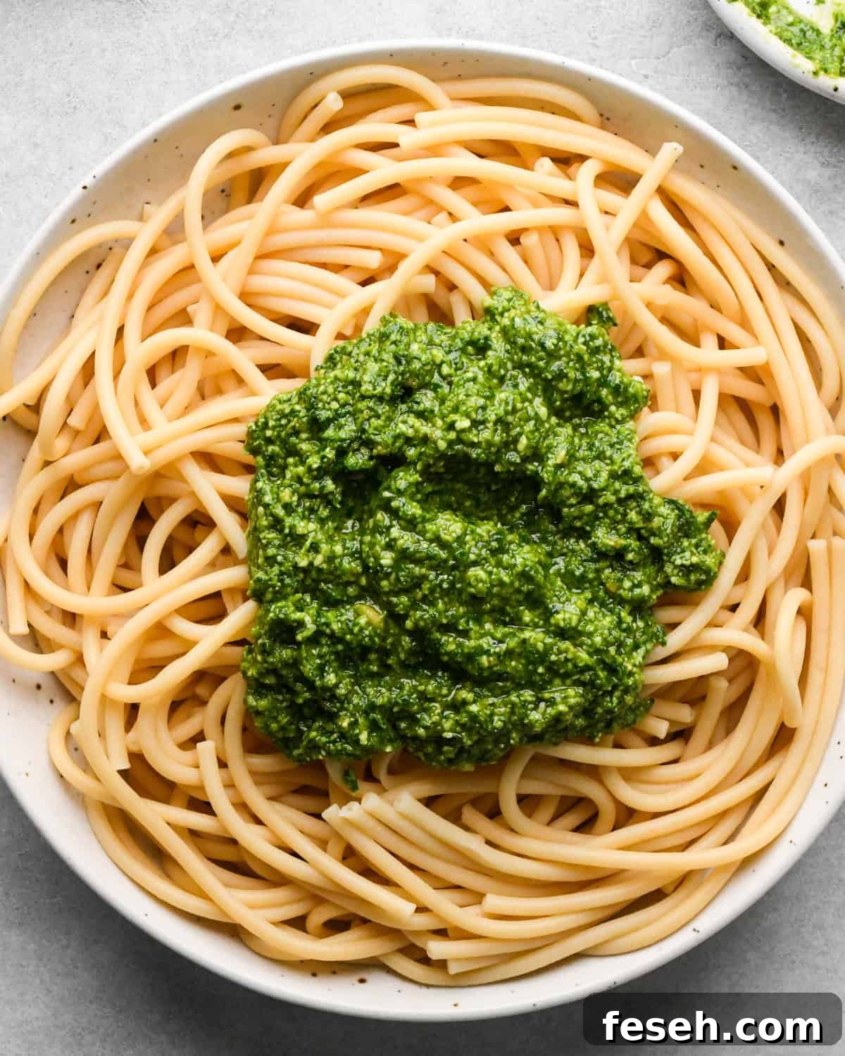 pesto sauce added on top of cooked pasta in a white bowl before mixing, making pesto pasta.
